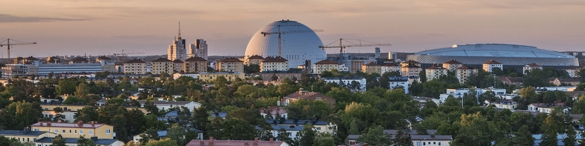 Stadsbild med bostadsområde i förgrunden och Globen i bakgrunden, omgiven av byggnader och byggkranar i kvällsljus